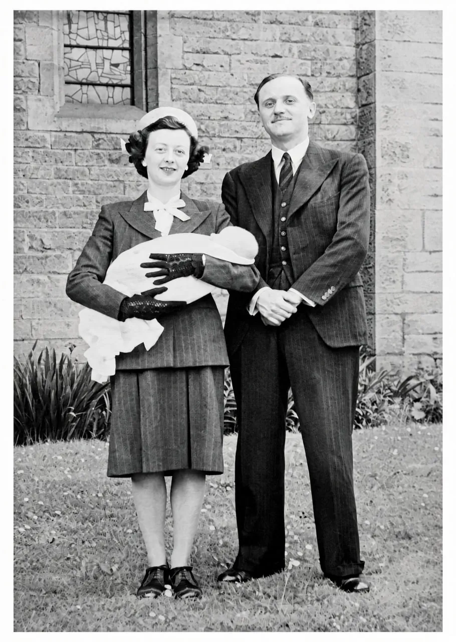 Black-and-white photograph of a man in a pinstripe suit and a woman in a suit and hat holding a baby wrapped in a white shawl, standing on a lawn in front of a stone church.