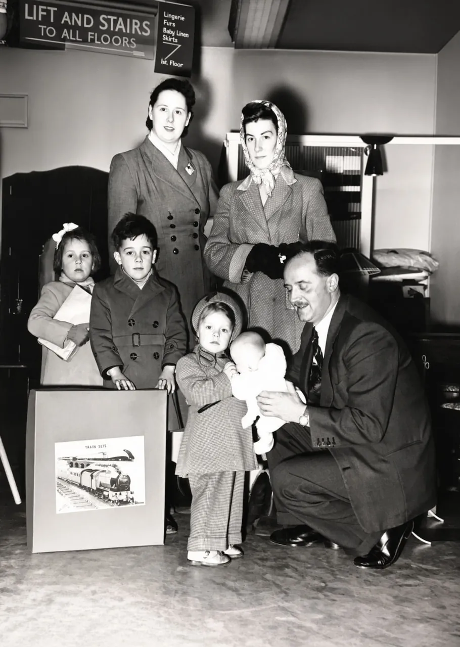 Black-and-white photograph inside a shop: a moustached man in a dark suit kneels beside a small child holding a baby doll, with two women and two more children standing behind and a boxed train set in front. Signs read 'Lift and stairs to all floors' and 'Lingerie, Furs, Baby Linen, Skirts, 1st Floor'.