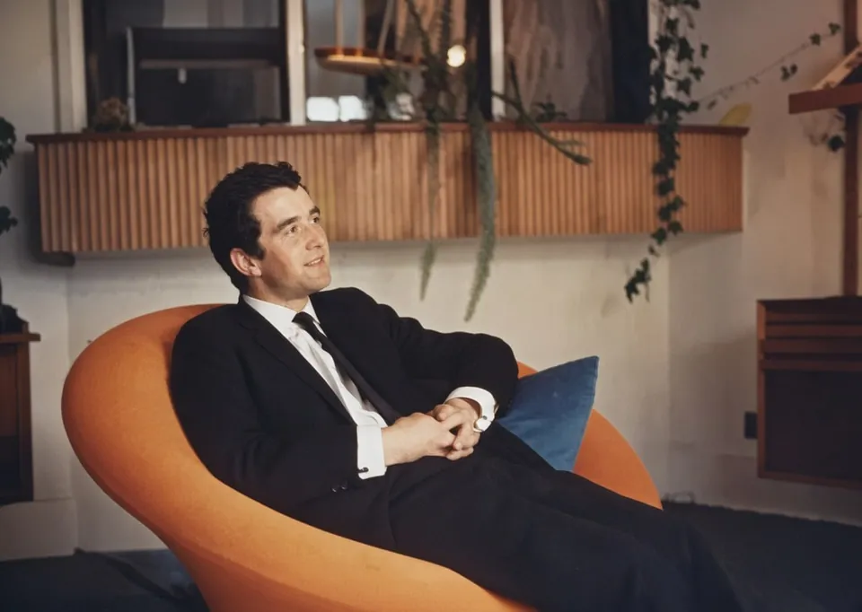 Colour photograph of a young man with dark hair, in a dark suit and tie, sitting back in a round orange armchair with a blue cushion, smiling, in a room with a slatted wooden shelf and trailing plants.