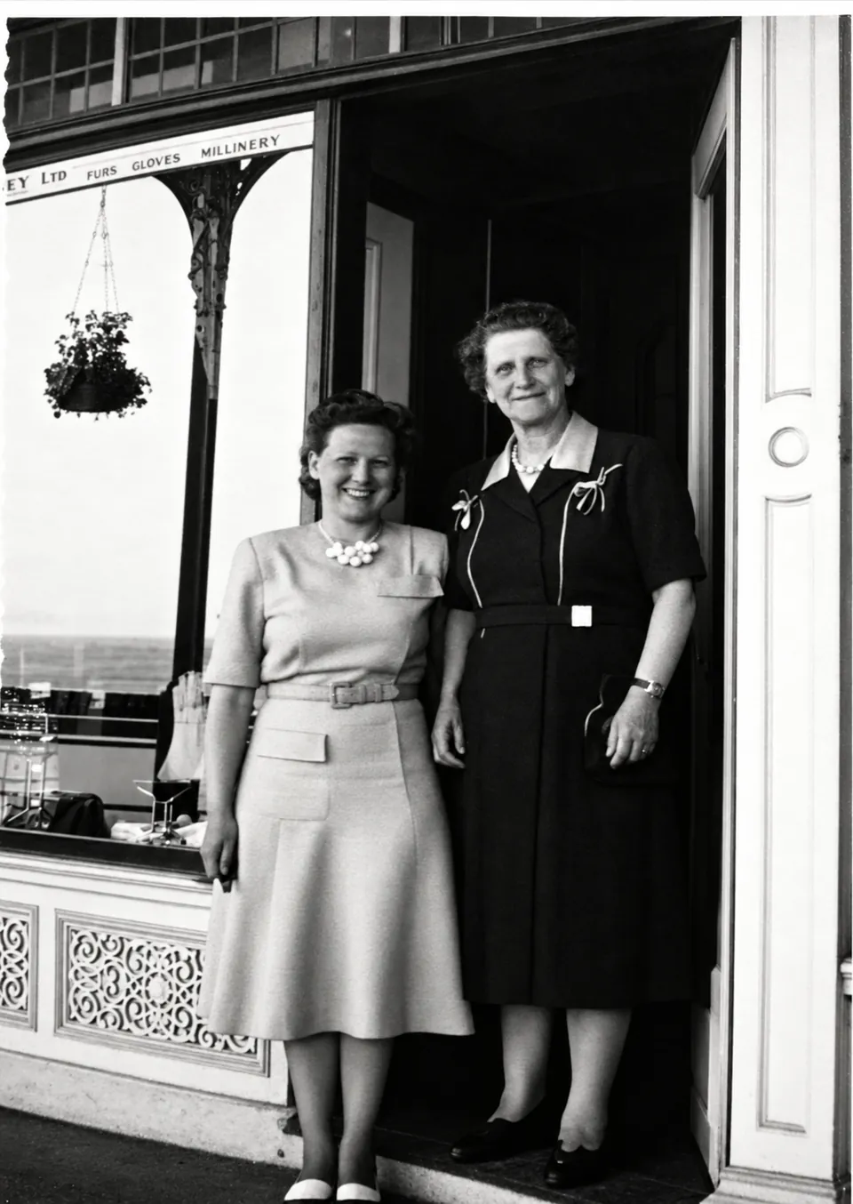 Black-and-white photograph of two women standing in a shop doorway, one in a light dress and one in a dark dress. The sign above the window reads '…ey Ltd. Furs Gloves Millinery'. A hanging flower basket is in the window.